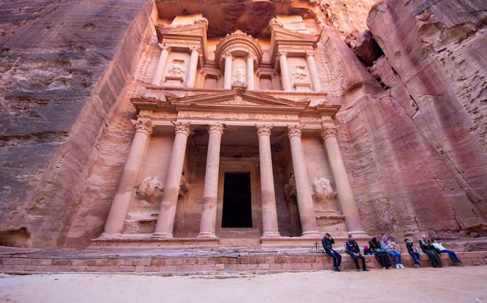 Petra's Al-Khazneh facade with visitors seated, Nabataean archaeological site, Jordan.