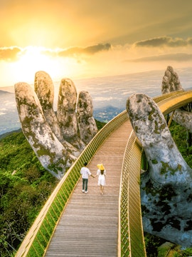 Couple walking on Golden Bridge at Sun World Ba Na Hills, Vietnam, with giant stone hands.