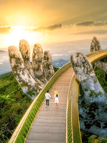 Couple walking on Golden Bridge at Sun World Ba Na Hills, Vietnam, with giant stone hands.