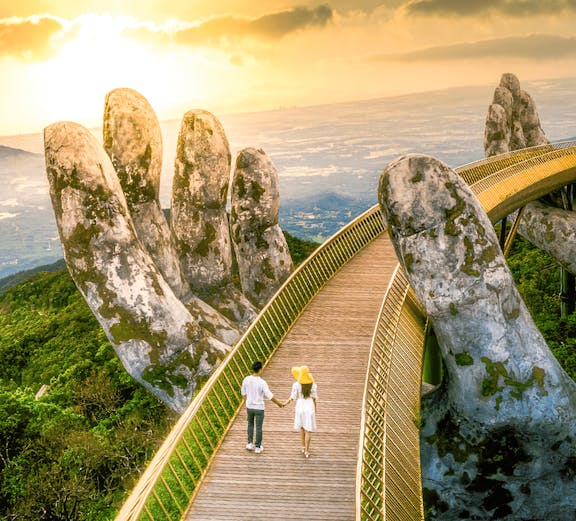 Couple walking on Golden Bridge at Sun World Ba Na Hills, Vietnam, with giant stone hands.
