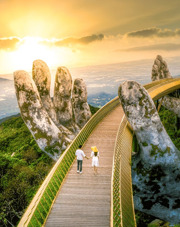 Couple walking on Golden Bridge at Sun World Ba Na Hills, Vietnam, with giant stone hands.