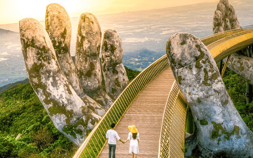 Couple walking on Golden Bridge at Sun World Ba Na Hills, Vietnam, with giant stone hands.