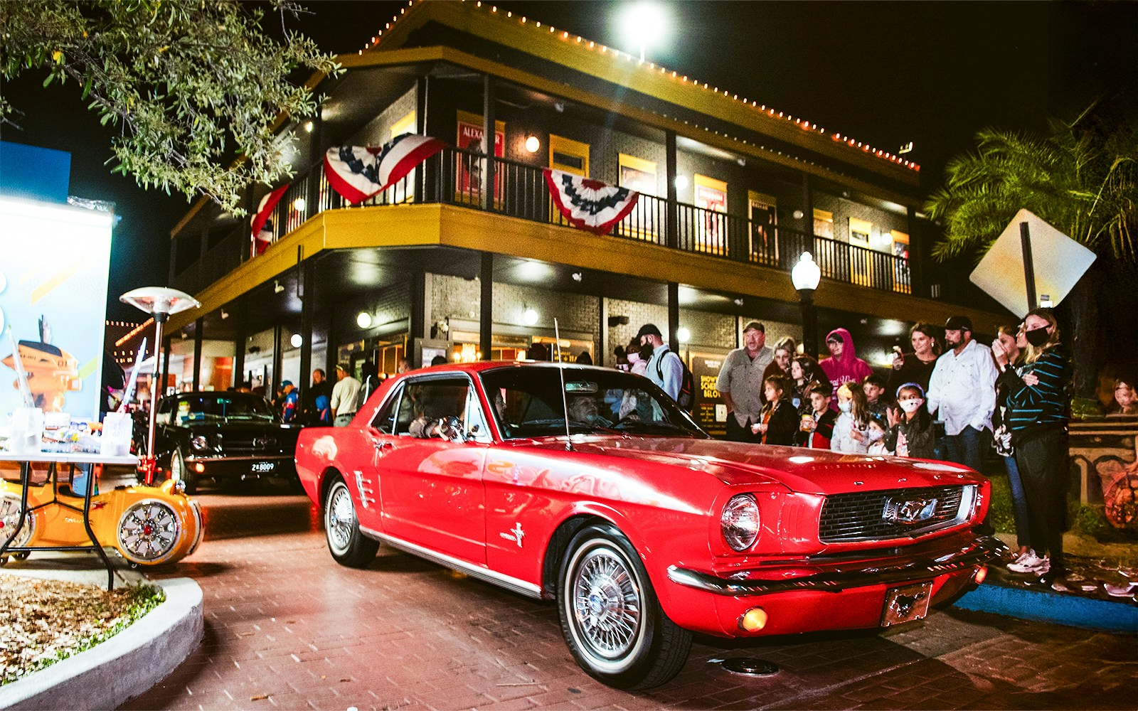 Red vintage car driving through Old Town, Orlando at night with a crowd watching.