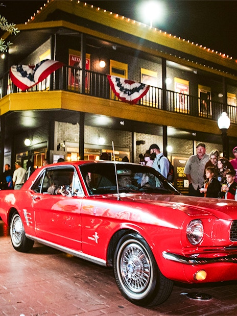 Red vintage car driving through Old Town, Orlando at night with a crowd watching.