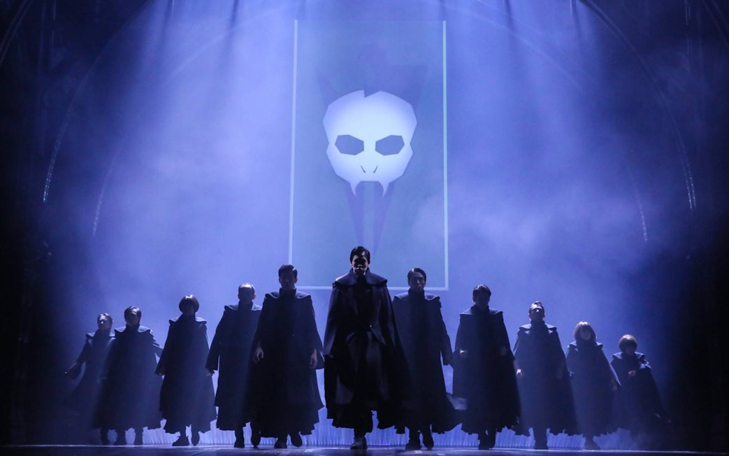 Cast performing on stage in "Harry Potter and the Cursed Child" with dark cloaks and a skull backdrop.