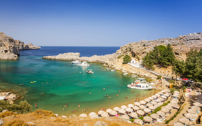 Beach in Lindos with turquoise water, boats, and sunbathers near rocky cliffs.