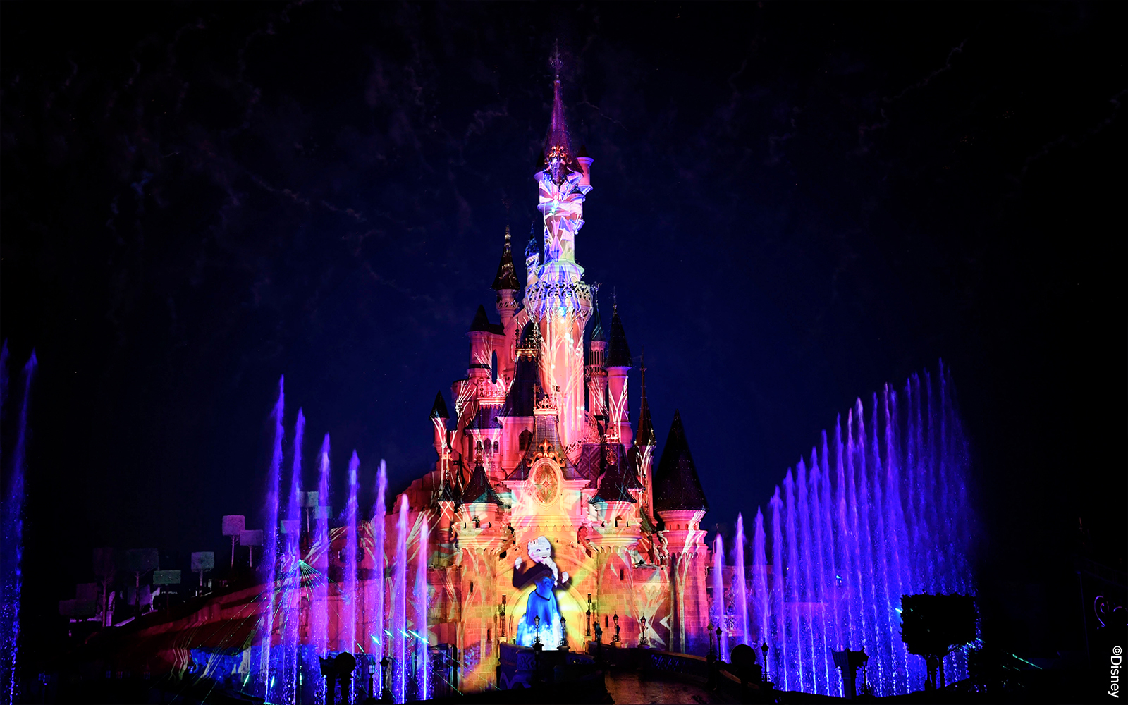 Disneyland Paris castle illuminated with colorful lights and fountains at night.