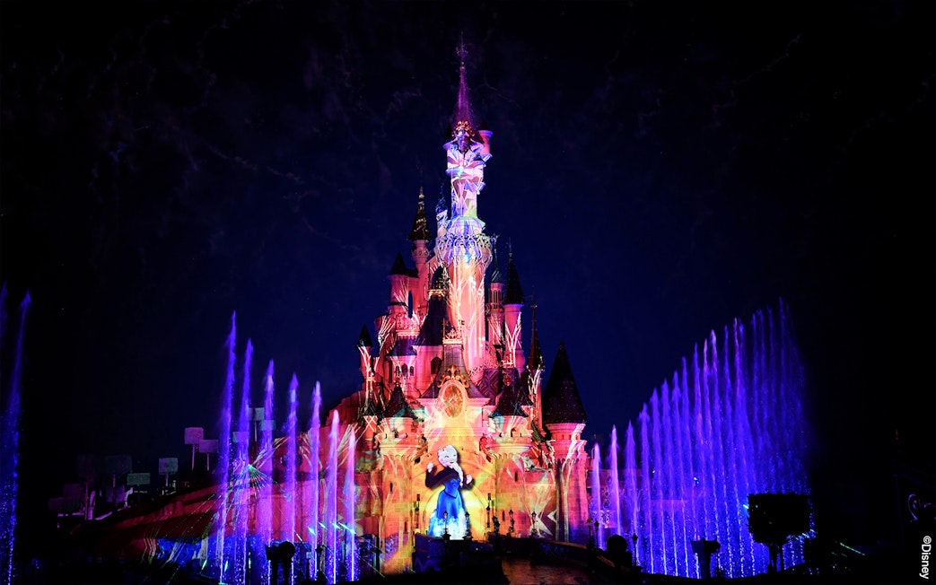Disneyland Paris castle illuminated with colorful lights and fountains at night.