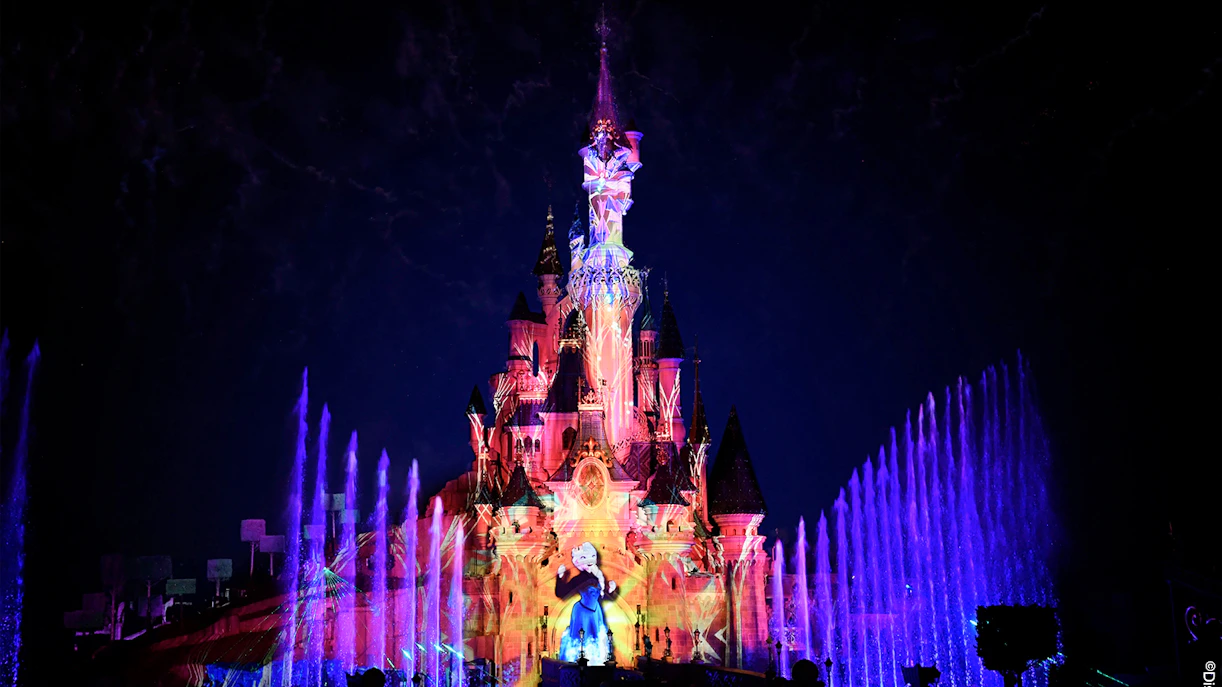 Disneyland Paris castle illuminated with colorful lights and fountains at night.