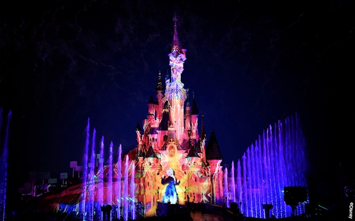 Disneyland Paris castle illuminated with colorful lights and fountains at night.