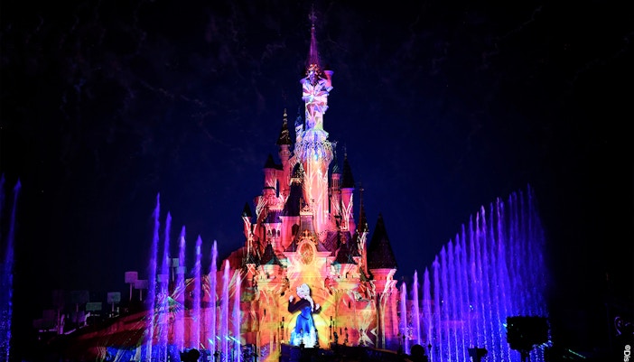 Disneyland Paris castle illuminated with colorful lights and fountains at night.
