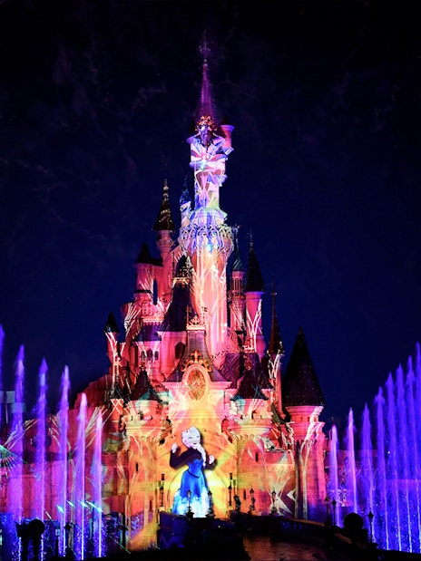 Disneyland Paris castle illuminated with colorful lights and fountains at night.