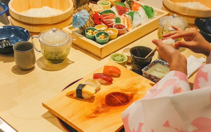 Tourist making sushi in traditional attire near Tokyo Tower with ingredients and tea set.