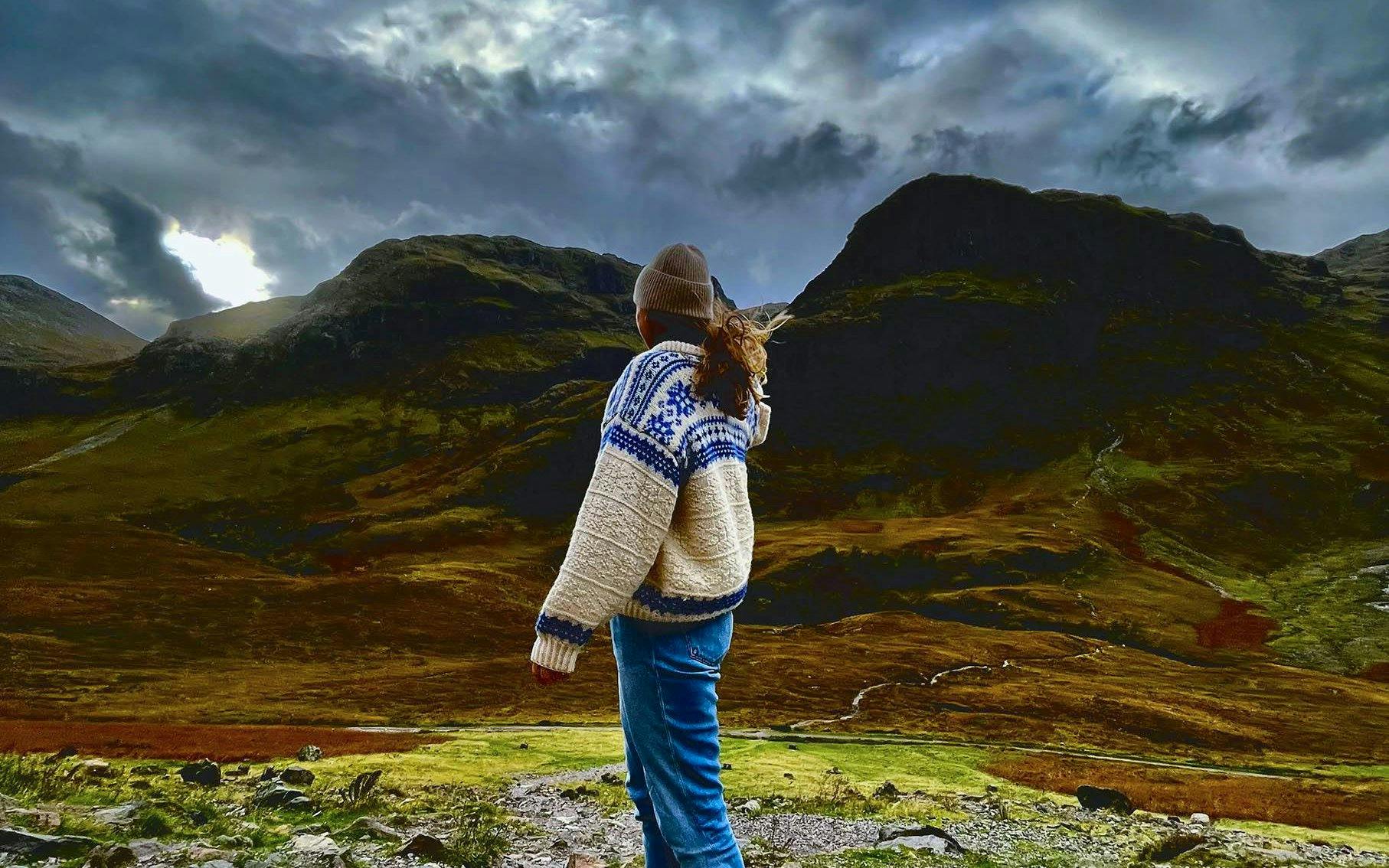 Person standing in Glencoe valley with dramatic mountain landscape.