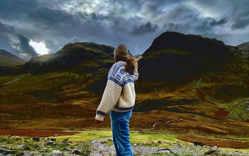 Person standing in Glencoe valley with dramatic mountain landscape.