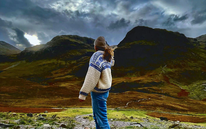 Person standing in Glencoe valley with dramatic mountain landscape.