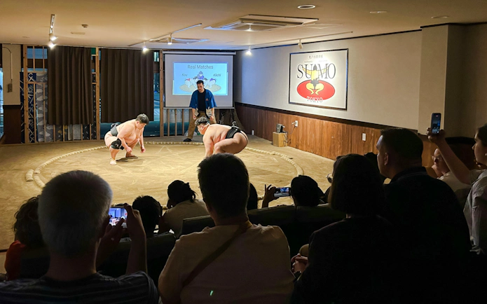 Sumo wrestlers preparing for a match in a Tokyo arena with an audience watching.