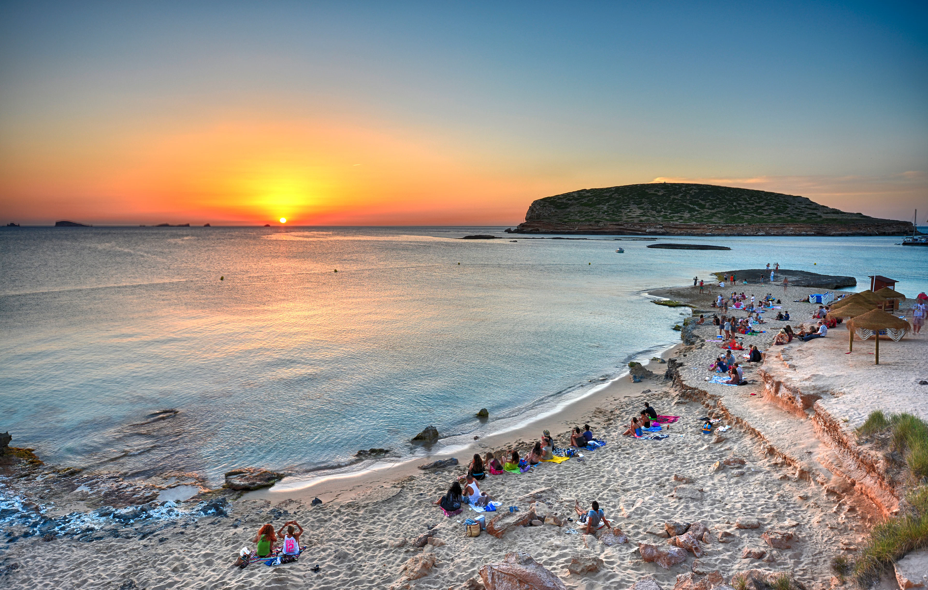 Sunset view over Cala Comte beach in Ibiza with people relaxing on the sand.