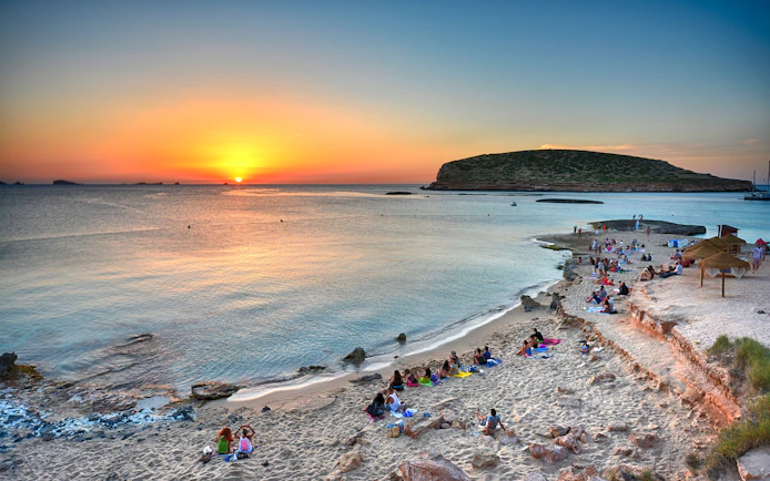 Sunset view over Cala Comte beach in Ibiza with people relaxing on the sand.