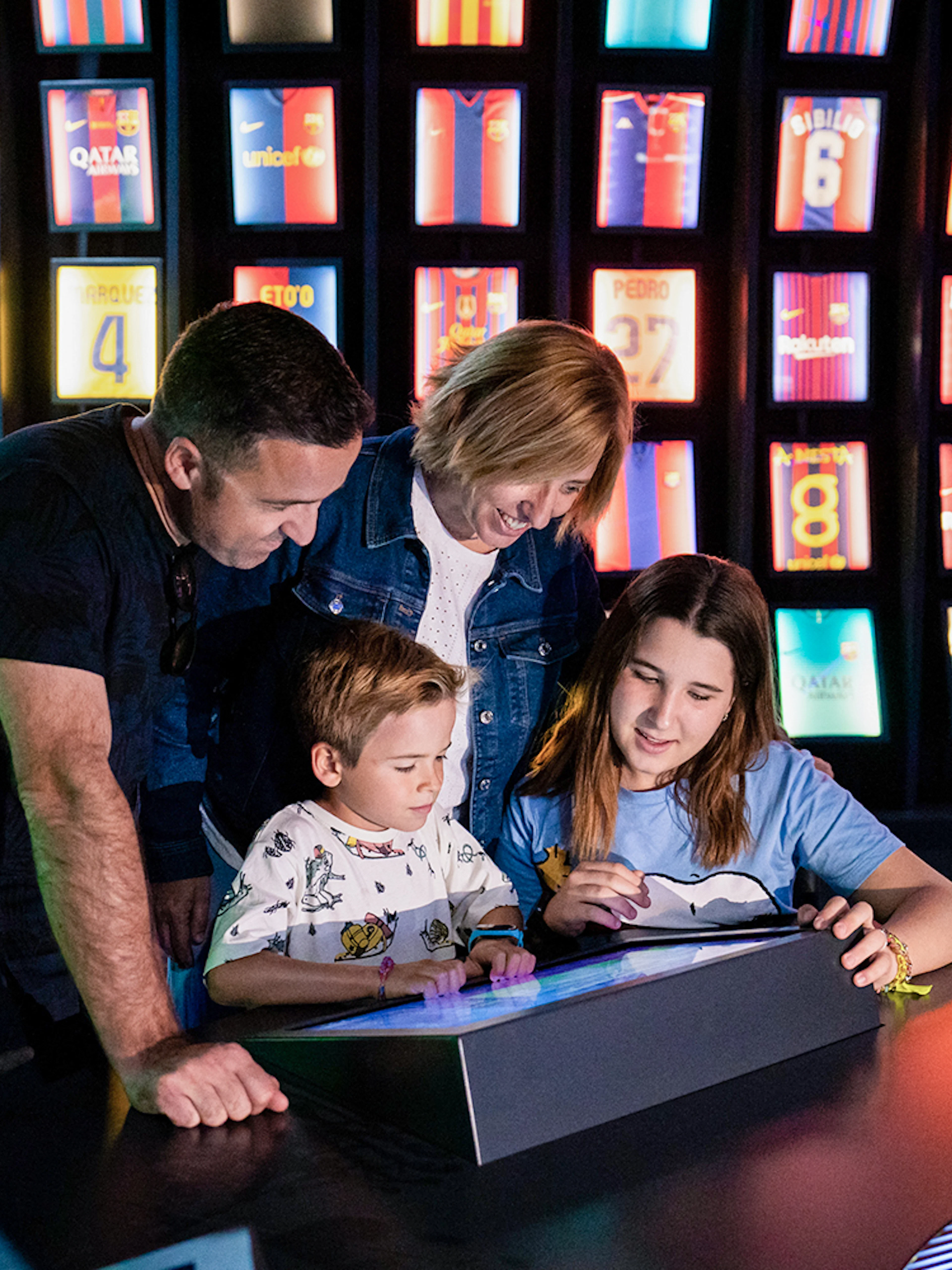 Family exploring interactive exhibit at Camp Nou Barca Museum with football jerseys in background.