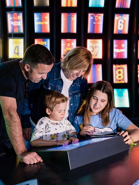 Family exploring interactive exhibit at Camp Nou Barca Museum with football jerseys in background.