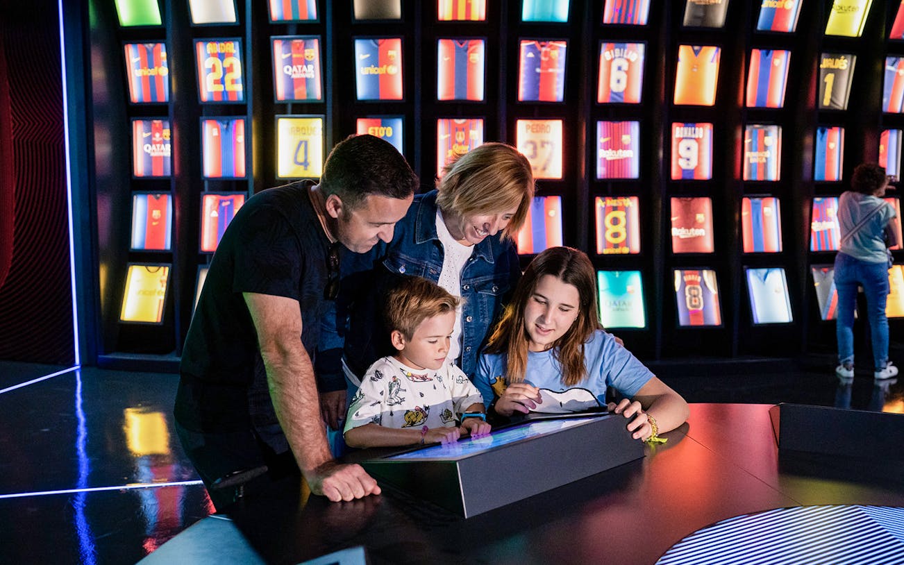 Family exploring interactive exhibit at Camp Nou Barca Museum with football jerseys in background.