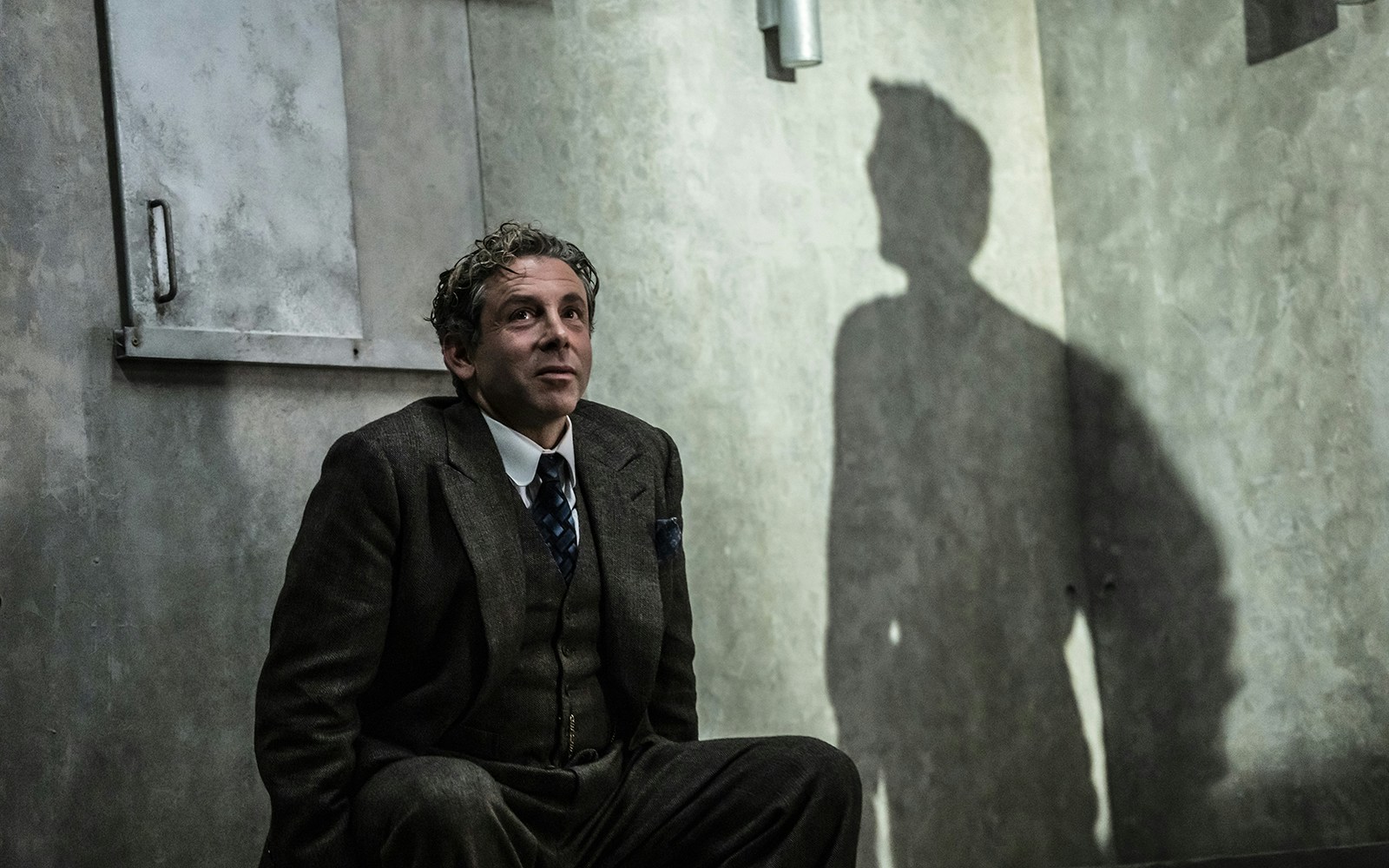 Man in a suit sitting against a wall with a shadow cast beside him.