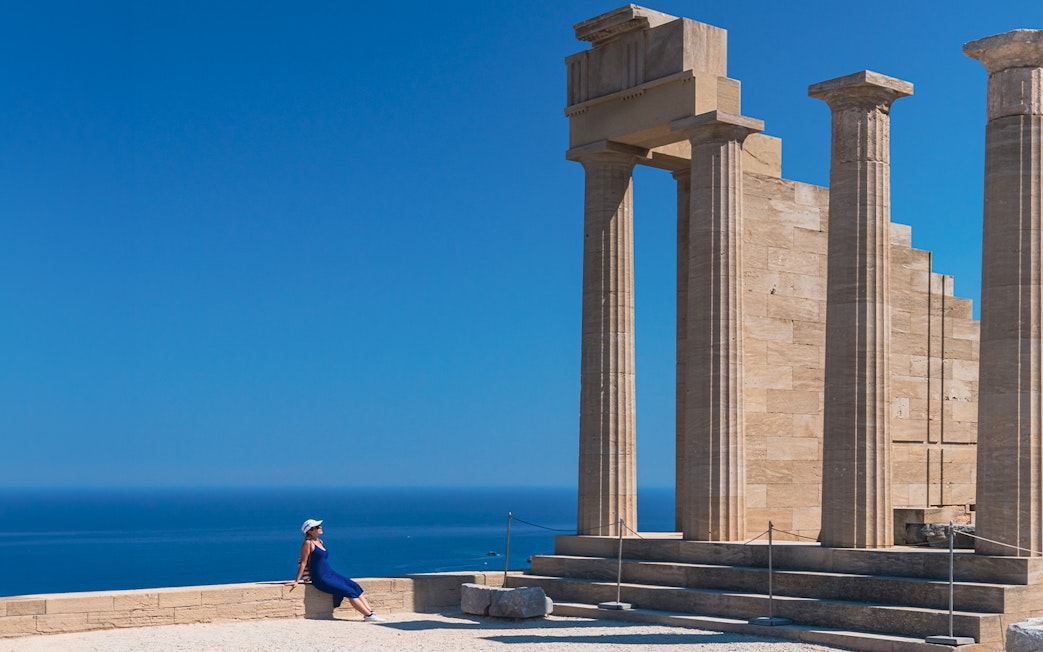 Ancient columns at Acropolis of Lindos with a person sitting nearby, overlooking the sea.