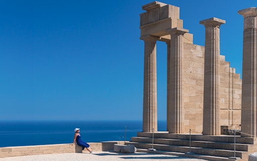 Ancient columns at Acropolis of Lindos with a person sitting nearby, overlooking the sea.