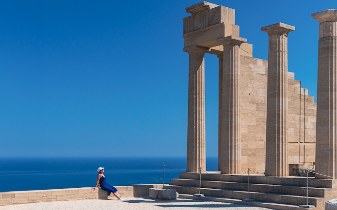 Ancient columns at Acropolis of Lindos with a person sitting nearby, overlooking the sea.