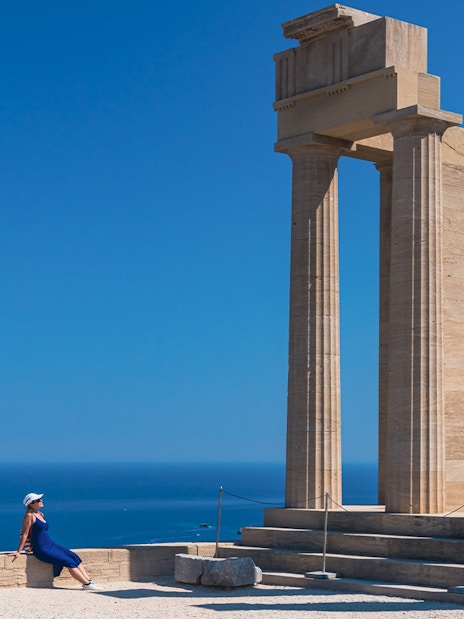 Ancient columns at Acropolis of Lindos with a person sitting nearby, overlooking the sea.