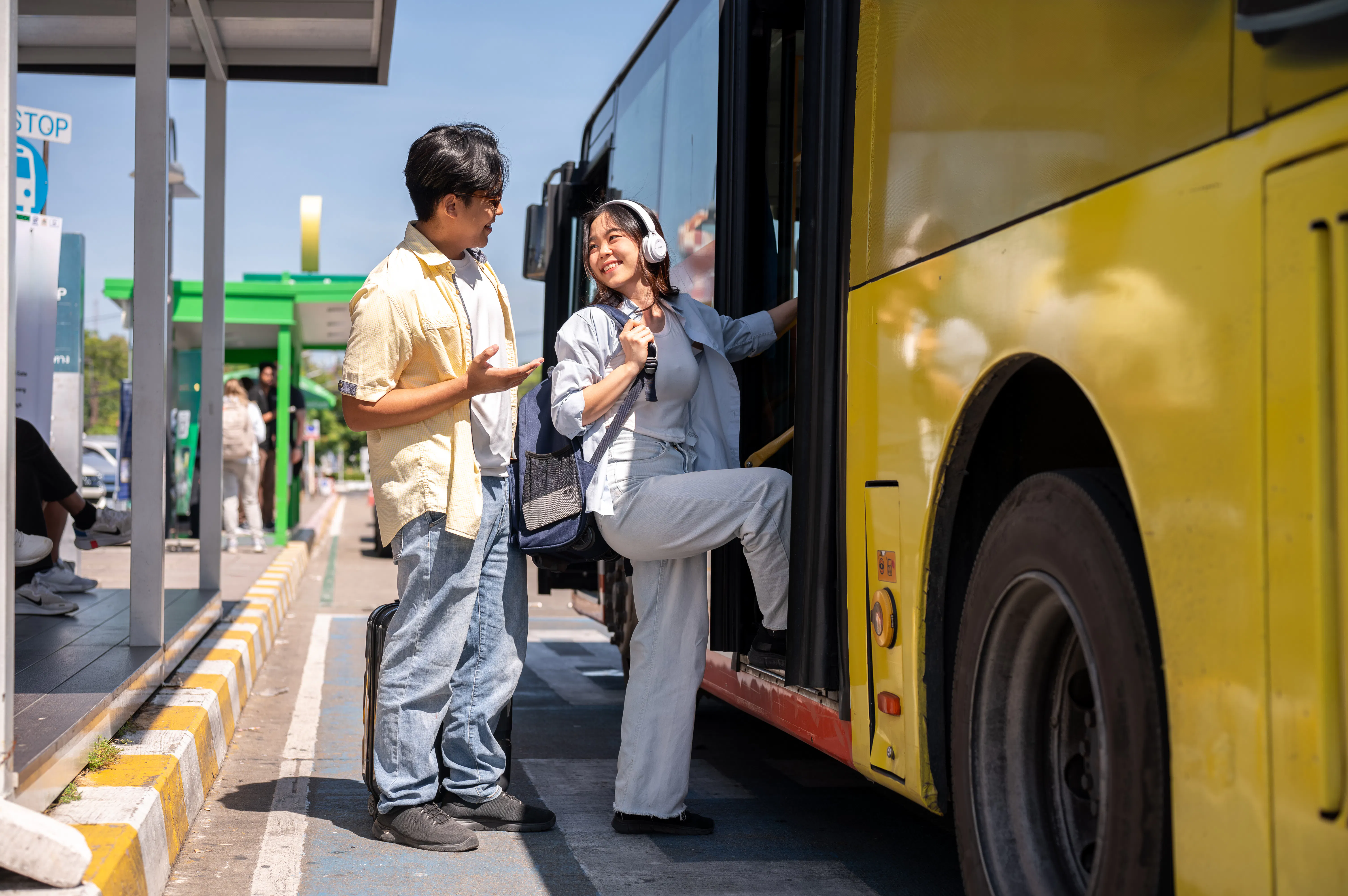 Tourist couple conversing at shuttle bus stop, woman boarding yellow bus.