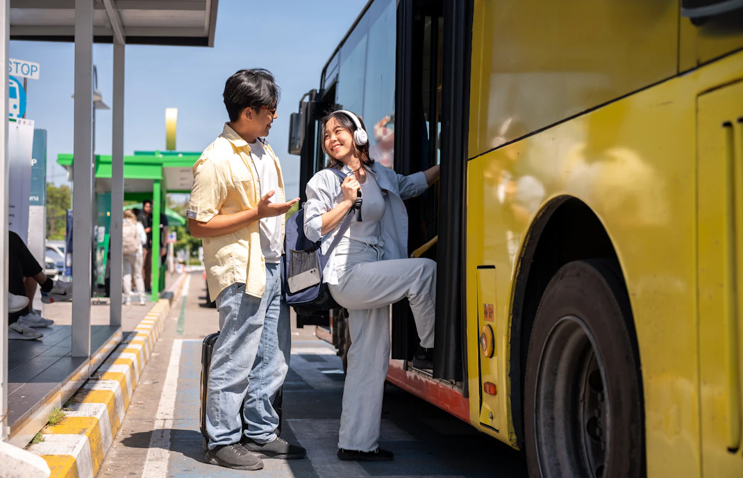 Tourist couple conversing at shuttle bus stop, woman boarding yellow bus.
