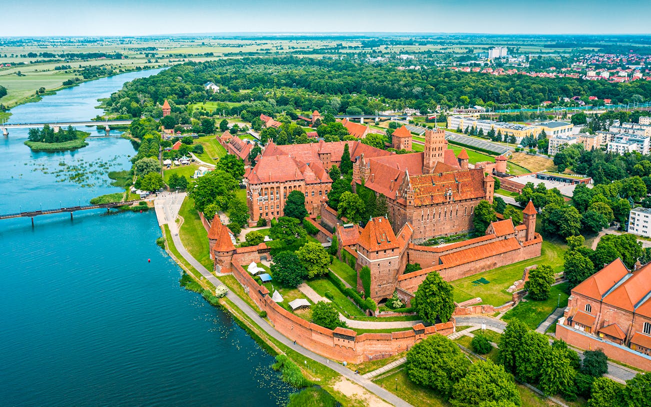 Aerial view of Malbork Castle surrounded by lush greenery and a river in Poland.