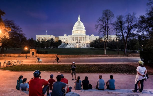 Tourists with bikes listen to a guide in front of the Capitol Building, Washington D.C. at night.