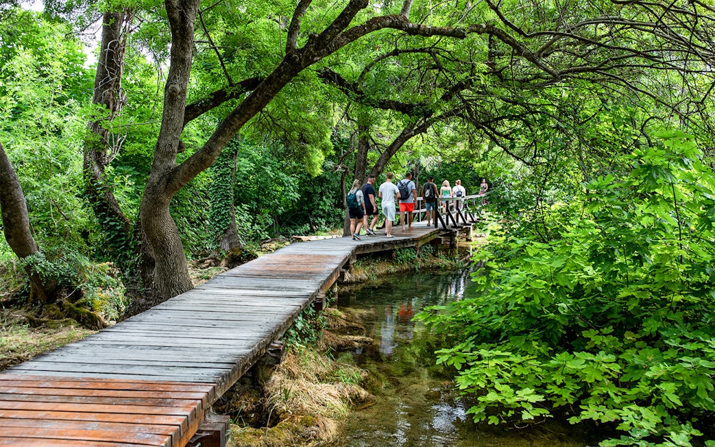 People walking on a wooden path through lush greenery in Krka National Park.