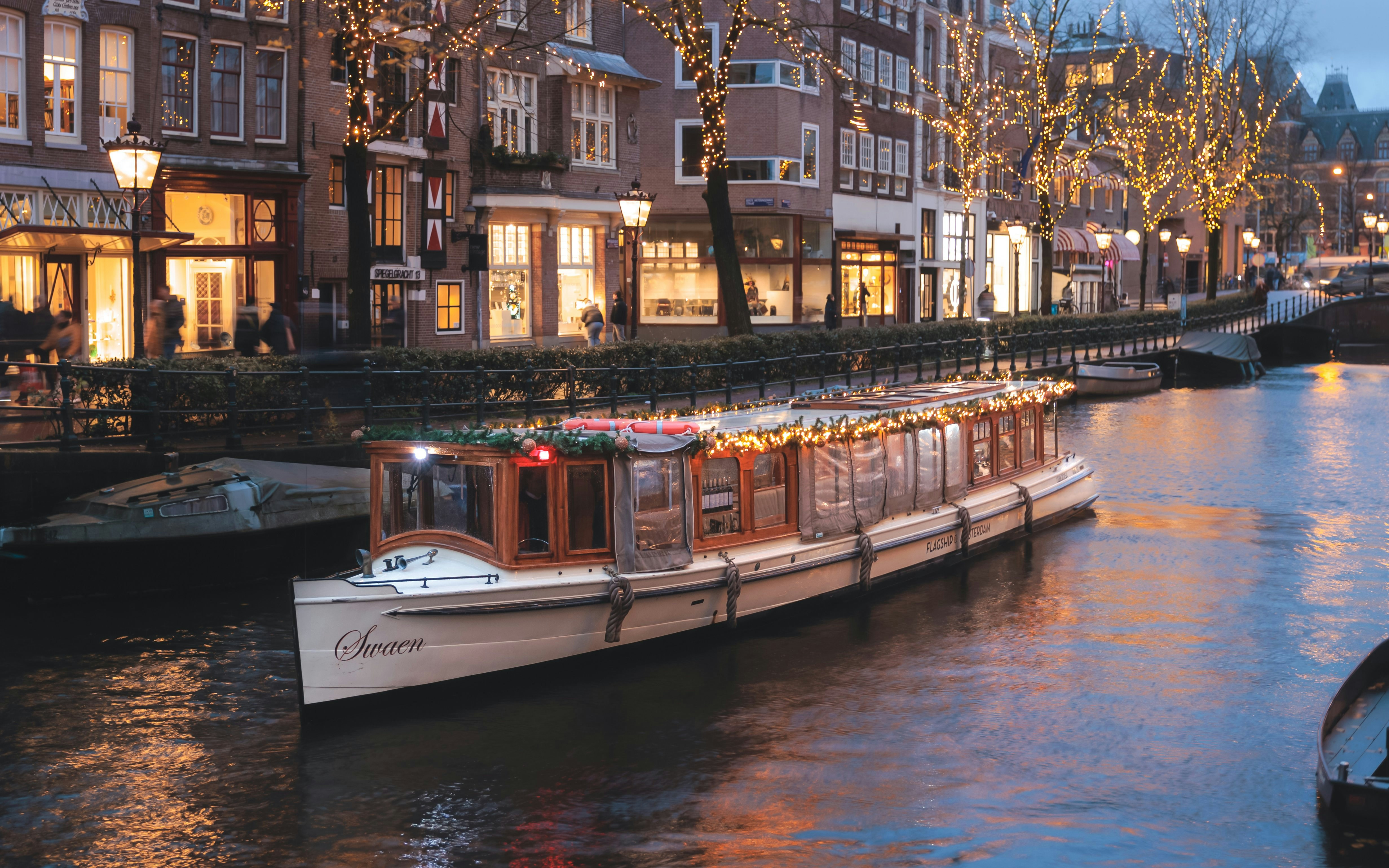 Luxury saloon boat adorned with lights cruising Amsterdam canal at dusk.