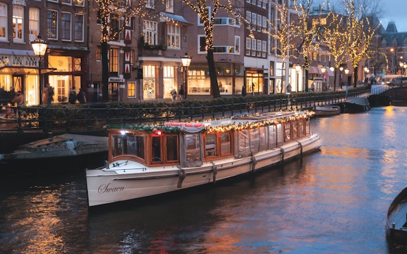 Luxury saloon boat adorned with lights cruising Amsterdam canal at dusk.