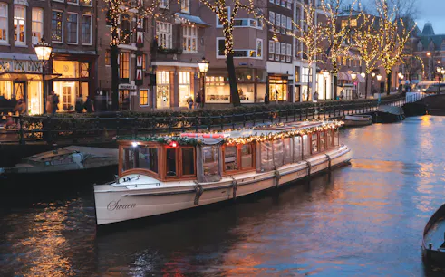 Luxury saloon boat adorned with lights cruising Amsterdam canal at dusk.