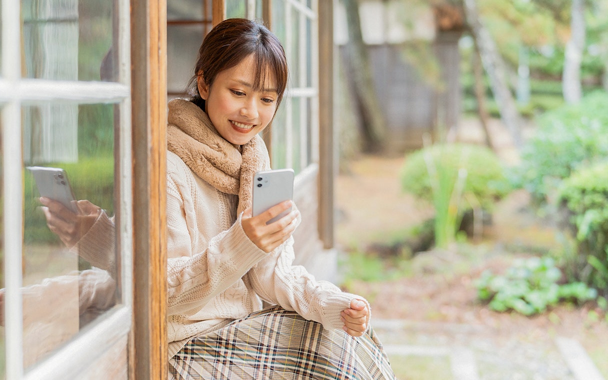 Woman sitting by a window, smiling while talking on a cellphone in a garden setting.