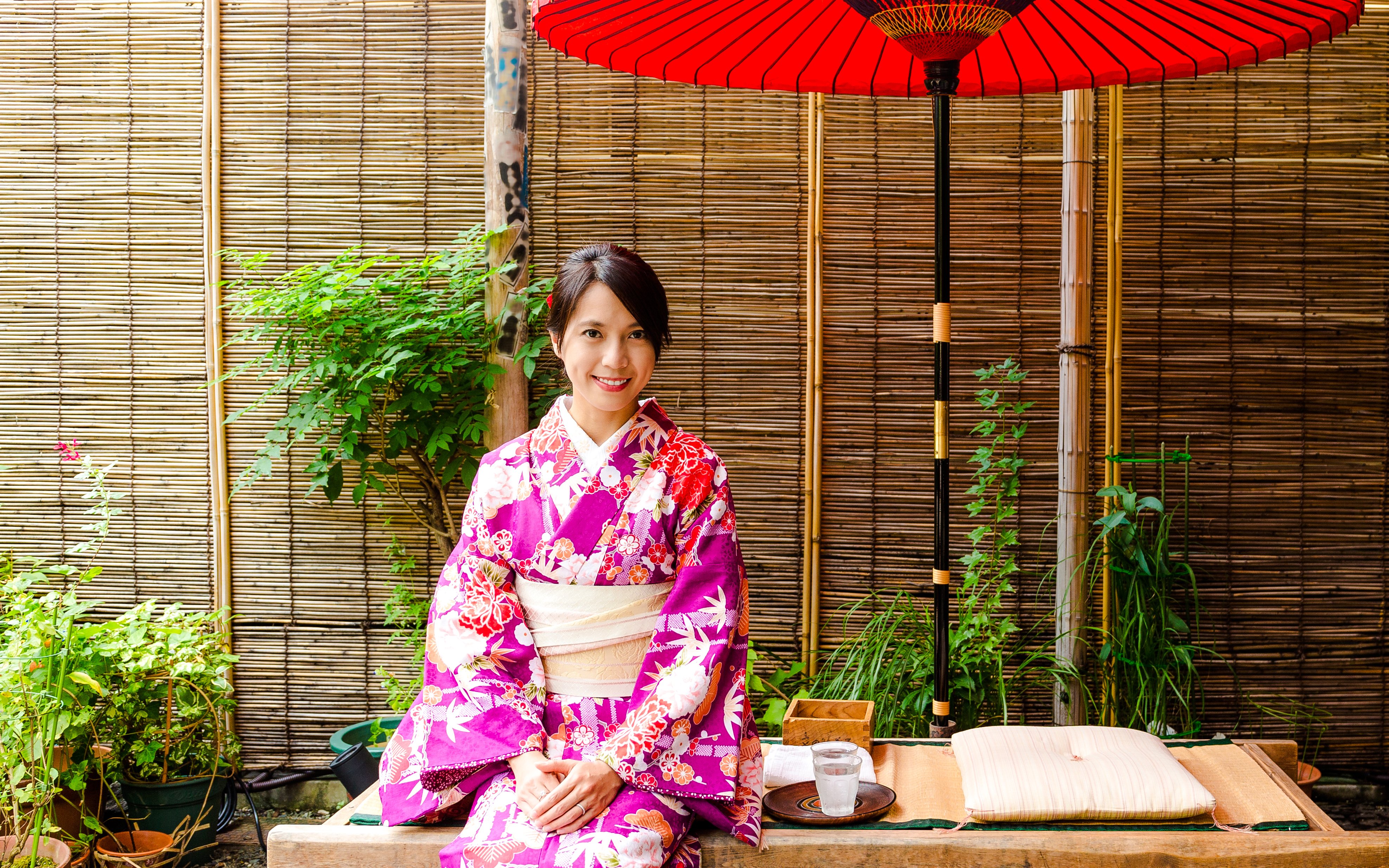 Japanese woman in kimono sitting at a traditional tea house with red umbrella.