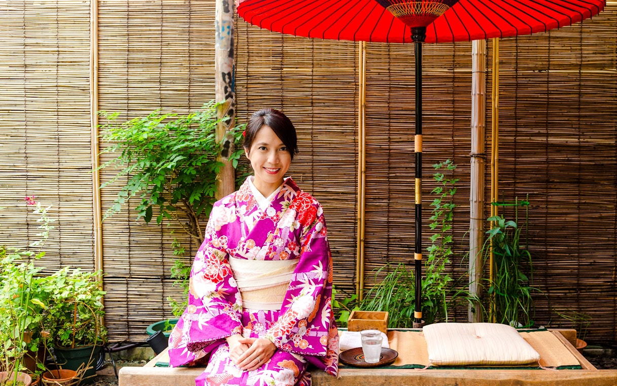 Japanese woman in kimono sitting at a traditional tea house with red umbrella.
