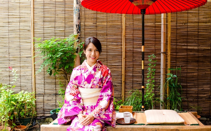 Japanese woman in kimono sitting at a traditional tea house with red umbrella.