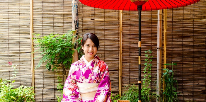 Japanese woman in kimono sitting at a traditional tea house with red umbrella.