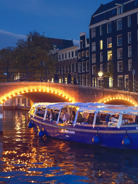 Covered canal cruise boat passing under illuminated bridge in Amsterdam at dusk.