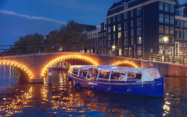 Covered canal cruise boat passing under illuminated bridge in Amsterdam at dusk.