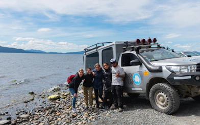 Group by 4x4 vehicle on rocky shore of Lake Fagnano during off-road tour.
