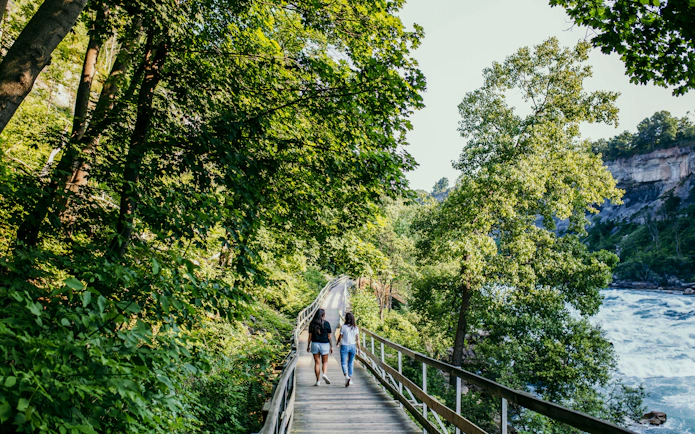 Visitors walking along a wooden path by the Niagara River surrounded by lush greenery.