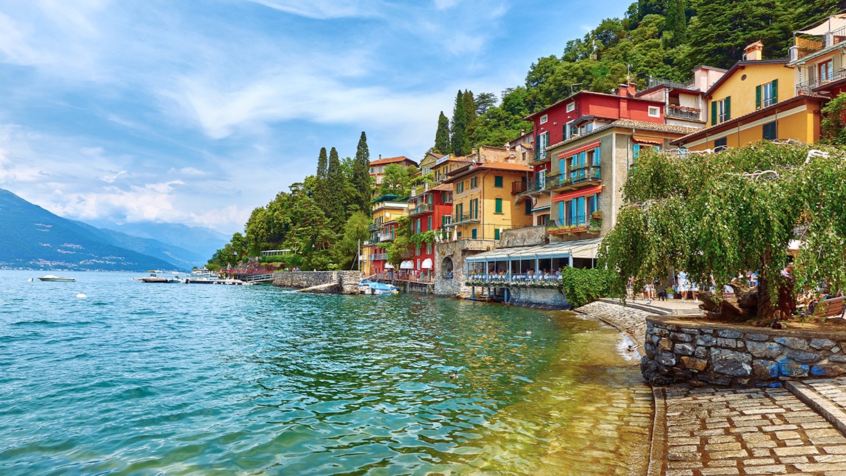 Colorful buildings along San Giovanni Beach, Lake Como, with clear water and lush greenery.