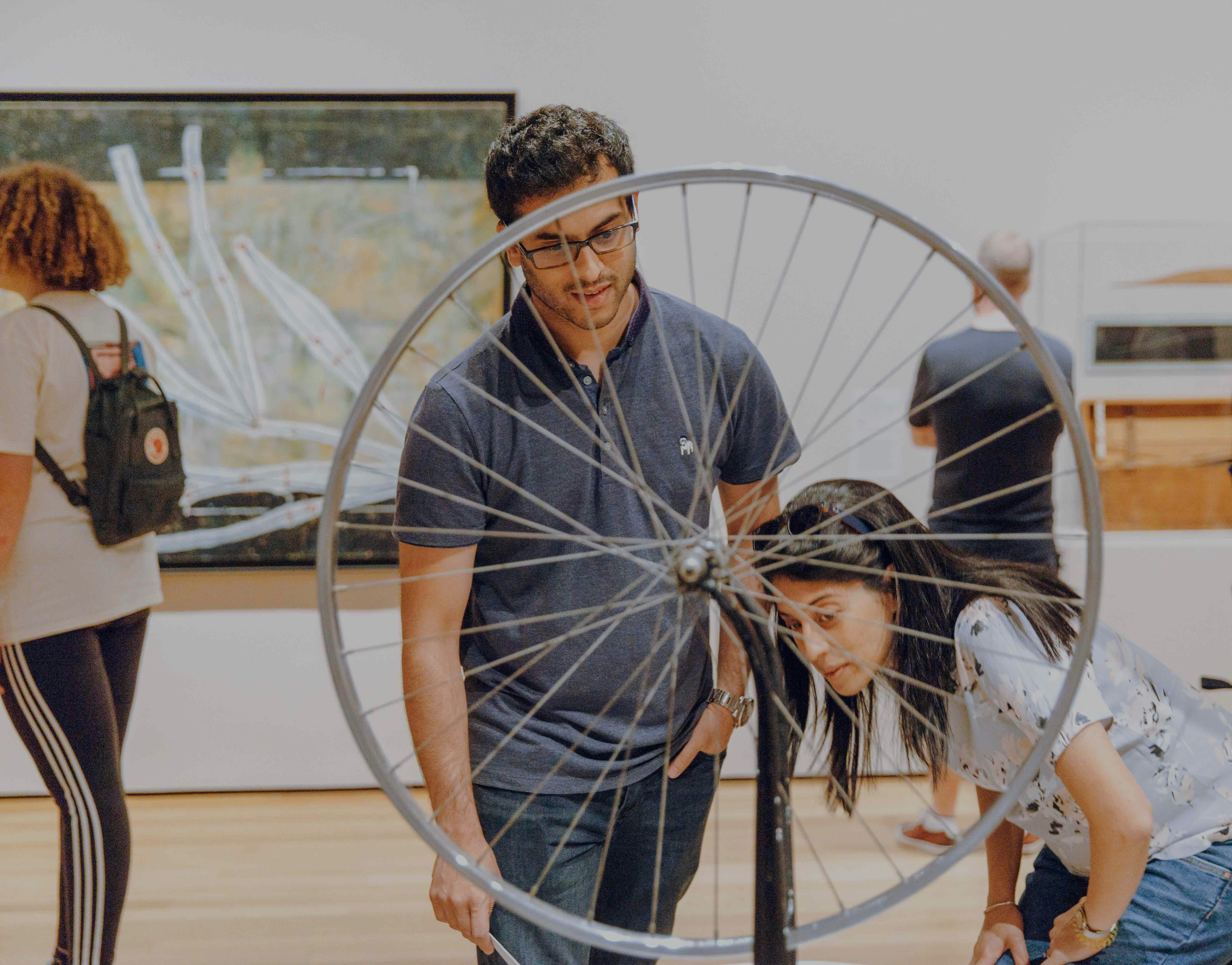 Visitors observing a bicycle wheel sculpture at NYC MoMA exhibition.